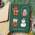 Load image into Gallery viewer, A dark green Christmas greeting card with four festive illustrations (gingerbread man, wrapped gifts, Santa in a red sweater, snowman) and the text 'Happy Holidays' at the bottom, styled with a kraft envelope, pine branches, and warm fairy lights.