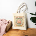 Load image into Gallery viewer, Tote bag with 'Pink Bunny Green Tea' design on a wooden surface with a plant in the background
