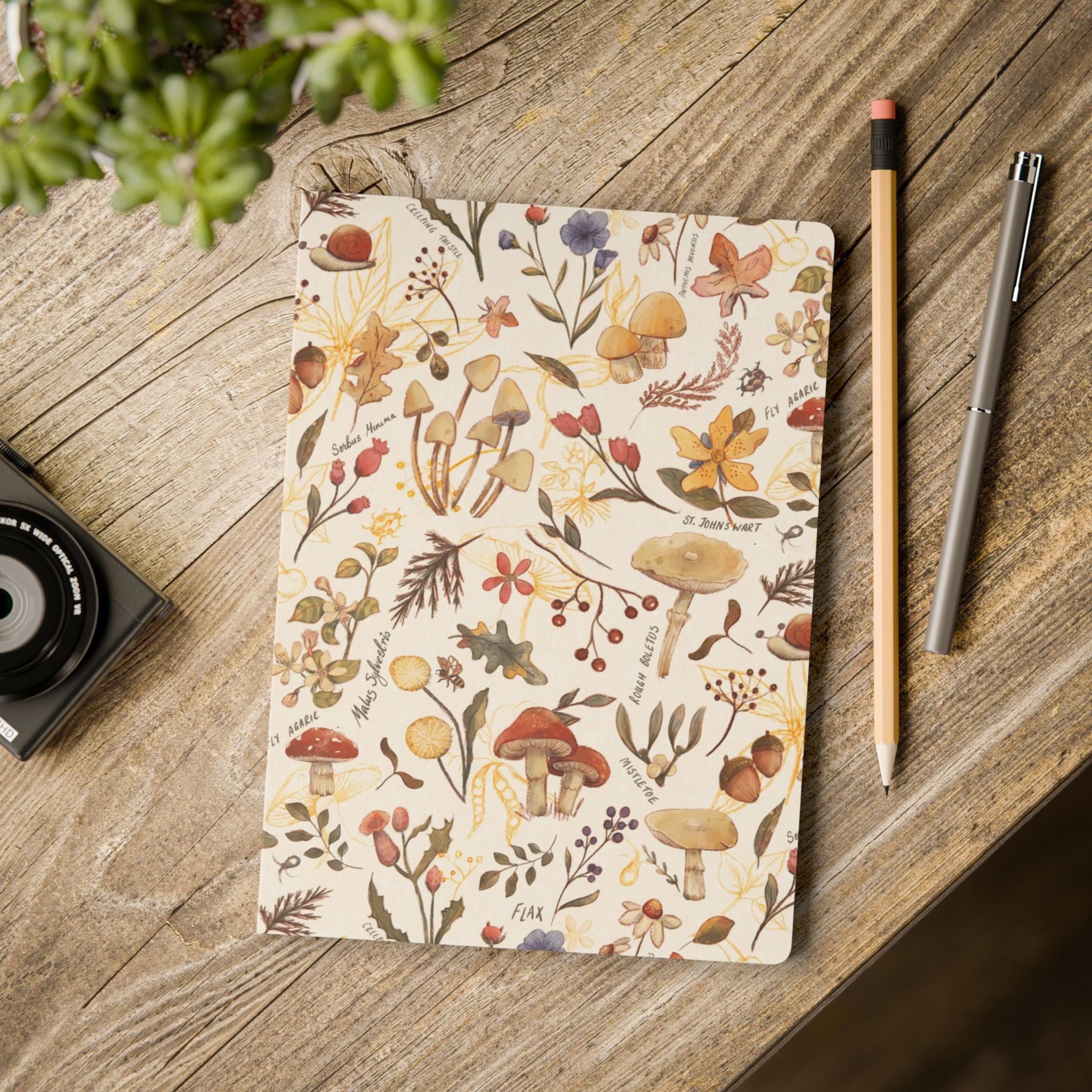 A flat-lay view of a notebook with a vintage-style botanical mushroom pattern on its cover, placed on a rustic wooden desk next to a pencil, a pen, and a camera.