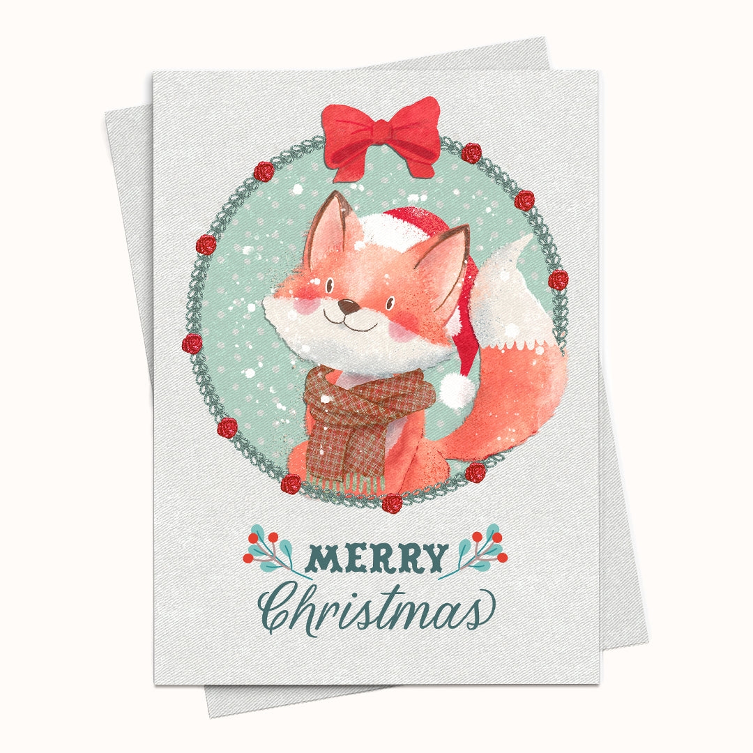 A Christmas greeting card featuring a watercolor illustration of a fox in a Santa hat and plaid sweater, framed by a holly wreath with a red bow, with 'MERRY Christmas' written below. The card is shown with a matching envelope on a light cream background.