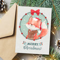 Load image into Gallery viewer, A Christmas greeting card featuring a watercolor illustration of a fox in a Santa hat and scarf inside a wreath, with the text 'Merry Christmas' below. The card is shown with a kraft envelope and holiday-themed flat-lay props including pine branches, fairy lights, and snowflakes.
