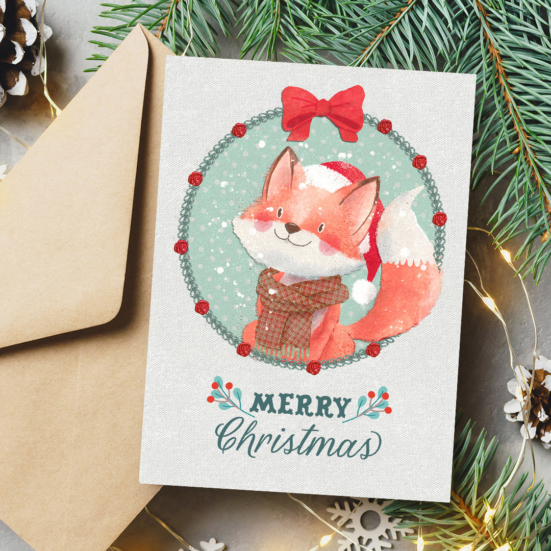 A Christmas greeting card featuring a watercolor illustration of a fox in a Santa hat and scarf inside a wreath, with the text 'Merry Christmas' below. The card is shown with a kraft envelope and holiday-themed flat-lay props including pine branches, fairy lights, and snowflakes.