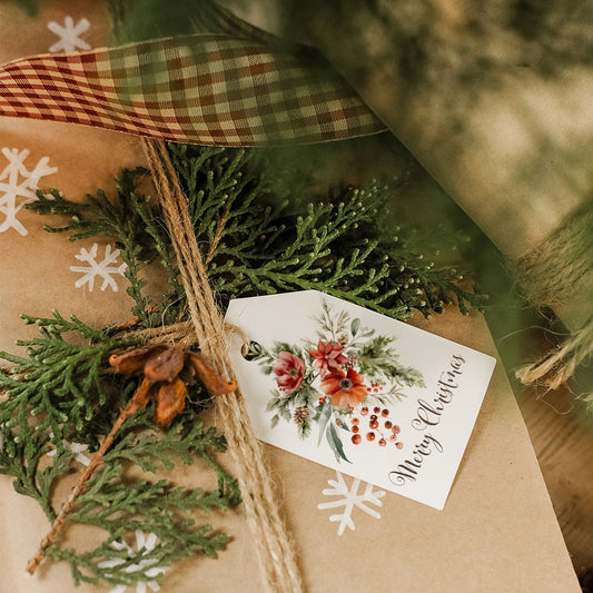 Gift wrapped in brown paper with greenery, a plaid ribbon, and a decorative tag on a wooden surface.