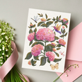 Load image into Gallery viewer, A watercolor-style floral art print or card featuring pink hydrangea blooms, green leaves, a hummingbird, butterflies, and dragonflies, styled on a light gray surface with a pink envelope and ribbon.
