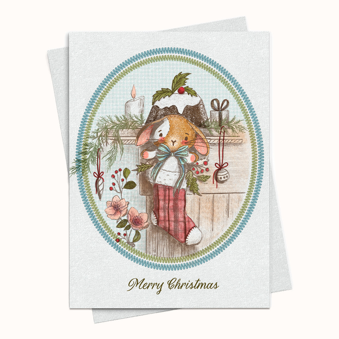 A Christmas greeting card with a vintage-style illustration of a rabbit in a stocking, surrounded by holiday decorations, with the text 'Merry Christmas' at the bottom. The card is shown with a matching envelope behind it on a light background.