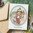 Load image into Gallery viewer, A Christmas greeting card featuring an illustration of a rabbit in a red plaid stocking, styled on a gray surface with a brown envelope, pine branches, and warm fairy lights.
