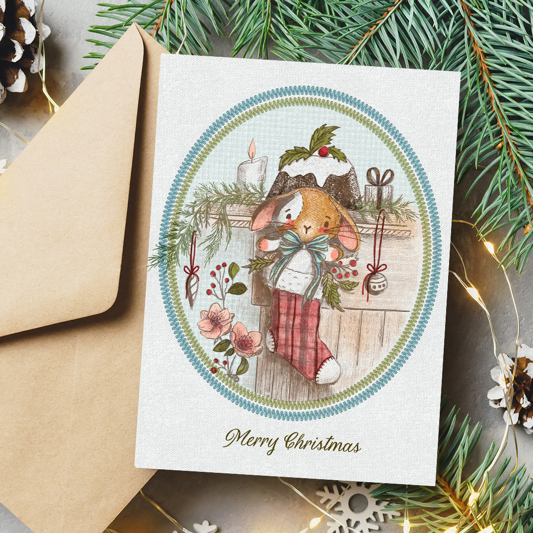 A Christmas greeting card featuring an illustration of a rabbit in a red plaid stocking, styled on a gray surface with a brown envelope, pine branches, and warm fairy lights.